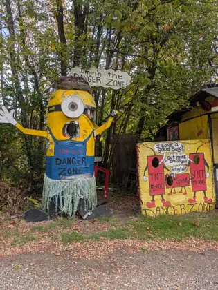 Roadside yard art: a wooden Minion figure in a grass skirt holds up a “Danger Zone” sign beside a hand-painted board covered in cartoon figures, under leafy trees.