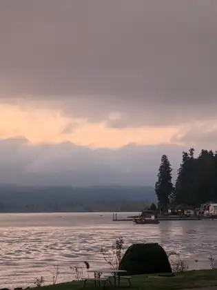 Dawn over the water at Potlatch: pink light glows beneath heavy grey clouds above a wooded point with houses and docks, with a large rock and a picnic table on the shore in front.