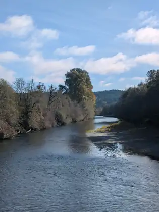 The river flows wide and shallow past gravel bars and banks of autumn trees, with forested hills beyond and white clouds in a blue sky.