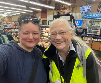 Jeana, in her yellow-green safety vest, and Amy, in a dark hoodie with sunglasses on her head, smile side by side in a selfie inside a minimart.