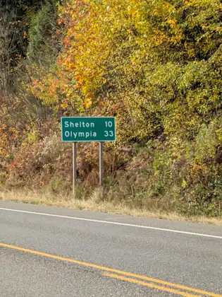 A green highway sign reading “Shelton 10, Olympia 33” stands on the roadside against a bank of trees in bright autumn yellow.
