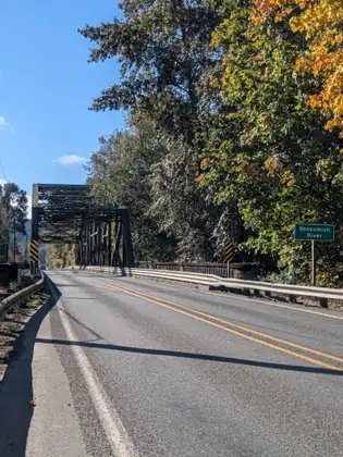 The highway crosses a steel truss bridge marked by a green “Skokomish River” sign, with trees in autumn colour on either side and a clear blue sky.