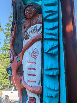 Close-up of a carved and painted totem pole at the trading post: a figure with a red face holds a white-painted form ringed in red, beside a column of carved bright blue shapes.