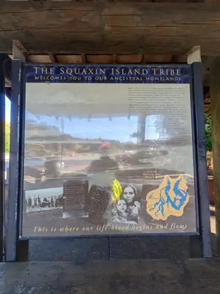 An interpretive sign under a timber shelter, headed “The Squaxin Island Tribe”, with historical photographs and a map of the inlets of southern Puget Sound.