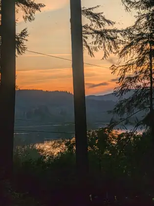 Sunset glows orange behind tall tree trunks and the outline of the hills, with a glint of the lake below.