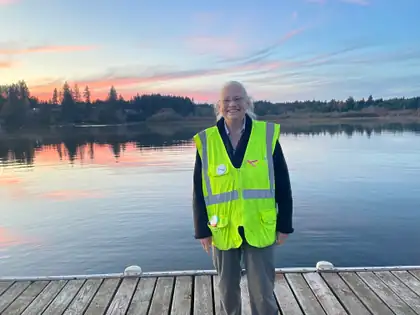 Jeana stands smiling on a wooden dock in her yellow-green safety vest, with the still lake behind her reflecting a pink and blue evening sky.