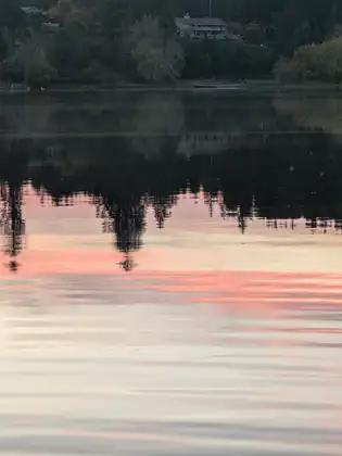 The trees and houses of the far shore reflected upside down in the lake, with pink sunset clouds rippling across the water.