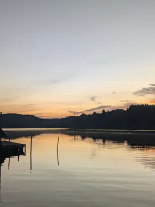 A small dock and a couple of posts stand in the still lake as the sky turns gold behind low wooded hills.