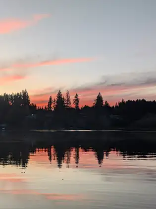 Tall firs stand in silhouette against a band of pink sunset on the far shore, doubled in the water below.