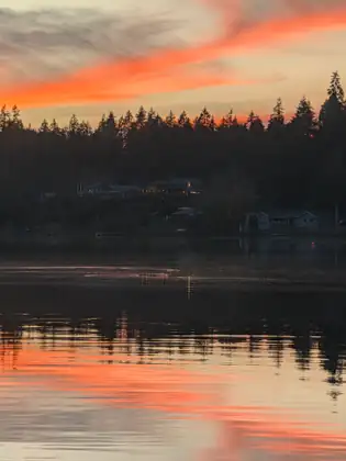 Bright orange clouds over the dark tree line and a few lit windows on the far shore, mirrored in the lake at dusk.