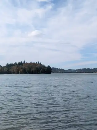 A wide stretch of grey-blue water, with a wooded point and a distant shore under soft white clouds.