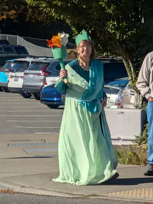 A woman dressed as the Statue of Liberty, in a long pale green gown and paper crown and holding up a torch, walks along the edge of a parking lot.