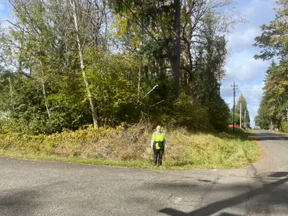 Jeana stands small on a grassy verge where a quiet road bends away into the trees.