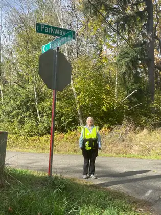 Jeana stands by a stop sign and street signs at a quiet road junction, with trees behind her.