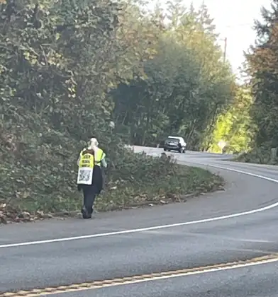 Seen from behind, Jeana walks the shoulder of a wooded bend in her yellow vest, with the QR code sign on her pack, as a car comes around the curve.