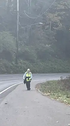 Jeana in her yellow vest walks toward the camera along the shoulder of a curving road beside a wooded hillside.