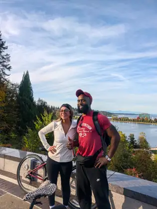 A woman in a white jacket and a man in a red shirt and cap stand beside a bicycle at a lookout above the water, with mountains in the distance.