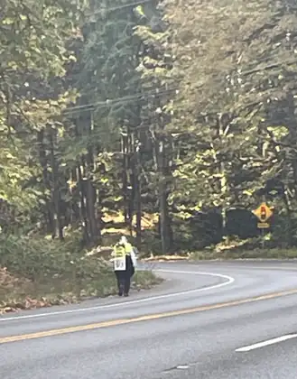 Far along a curving, tree-lined road, Jeana walks the shoulder toward the camera in her yellow vest.