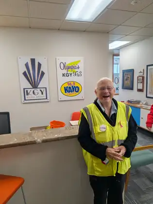 Jeana, laughing, stands at a reception counter in her yellow safety vest, with KGY Media Group, Olympia’s KGY 95.3 and 96.9 KAYO Country signs on the wall behind her.