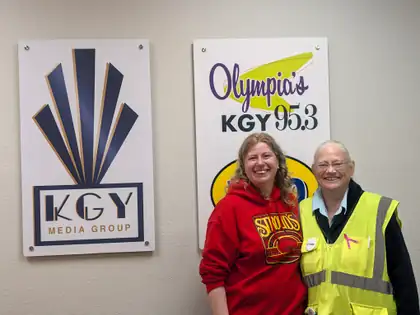 Jeana in her yellow safety vest stands beside a smiling woman in a red hooded sweatshirt, in front of KGY Media Group and Olympia’s KGY 95.3 signs.