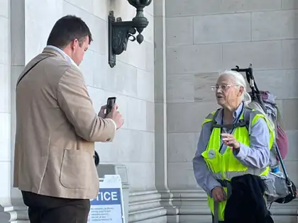 Jeana talks outside the Capitol while a man in a tan jacket holds up a phone.