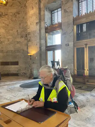 Still wearing her backpack and safety vest, Jeana writes in an open book on a table inside the Capitol.
