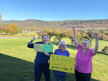 Jeana’s sister Ramona and two friends cheer in a sunny field above an autumn valley, holding hand-lettered signs reading “Hurray Jeana! Steps to Marrow V” and “Bravo! Seattle to Vancouver, Canada to Olympia, WA. Jeana, 315 miles, is awesome!”