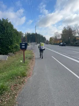 Jeana, wearing her yellow safety vest and backpack, walks along a paved roadside path beneath a blue sky.