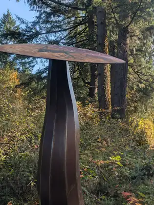 A tall metal sculpture shaped like a mushroom stands beside a trail among autumn trees.