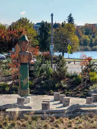 A carved and painted wooden figure in a green hat stands in a landscaped waterside park, with short wooden posts set in the ground around it.