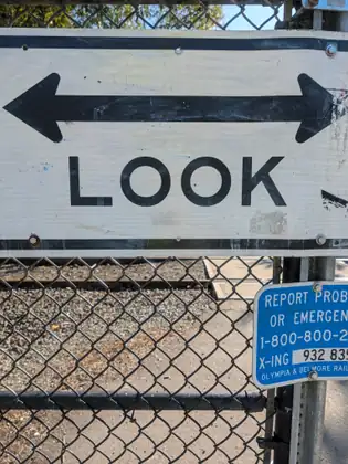 A railway crossing sign with a double-headed arrow reads LOOK, fixed to a chain-link fence.