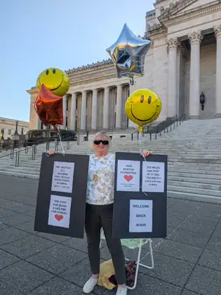 A woman holds up boards of signs for Jeana at the foot of the Capitol steps, with smiley-face and star balloons above her.