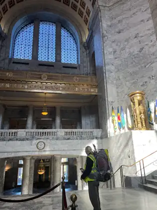 Seen from behind in her backpack and vest, Jeana looks around the Capitol rotunda, with flags, a gilded balcony and a tall arched window above.