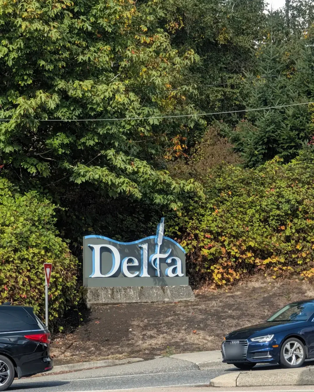The Delta city sign, white letters edged in blue on a grey panel set on a stone base, in front of trees turning orange.