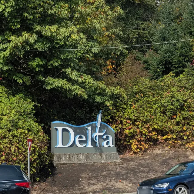 The Delta city sign, white letters edged in blue on a grey panel set on a stone base, in front of trees turning orange.